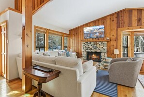 Ein gemütliches Wohnzimmer mit Kamin aus Stein, Holzwänden und einem großen Fenster mit Blick auf eine verschneite Landschaft.