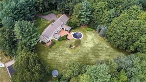 An aerial view of a two-story house with a large front yard, surrounded by dense trees. - Spacious 5-bedroom house in gorgeous, convenient Avon with pool and fitness room (Avon)