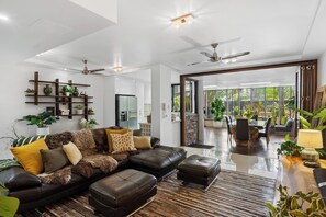 A modern living room with a leather sofa, a dining area with a wooden table, and a view of a garden through large windows. - Luxury Beachfront Apartment with Direct Pool Access (Cairns)