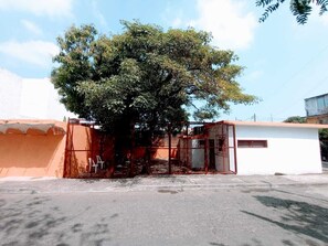 Un edificio con entrada protegida por una cerca, una estructura pequeña contigua y un árbol grande al frente.