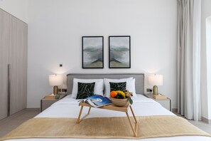 A modern hotel room with a large bed, two bedside tables with lamps, a bedside table with a fruit bowl, and two framed abstract paintings on the wall.