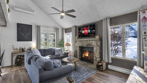 A modern living room with a fireplace, a large TV displaying the Netflix logo, and a view of a snowy landscape through the windows.