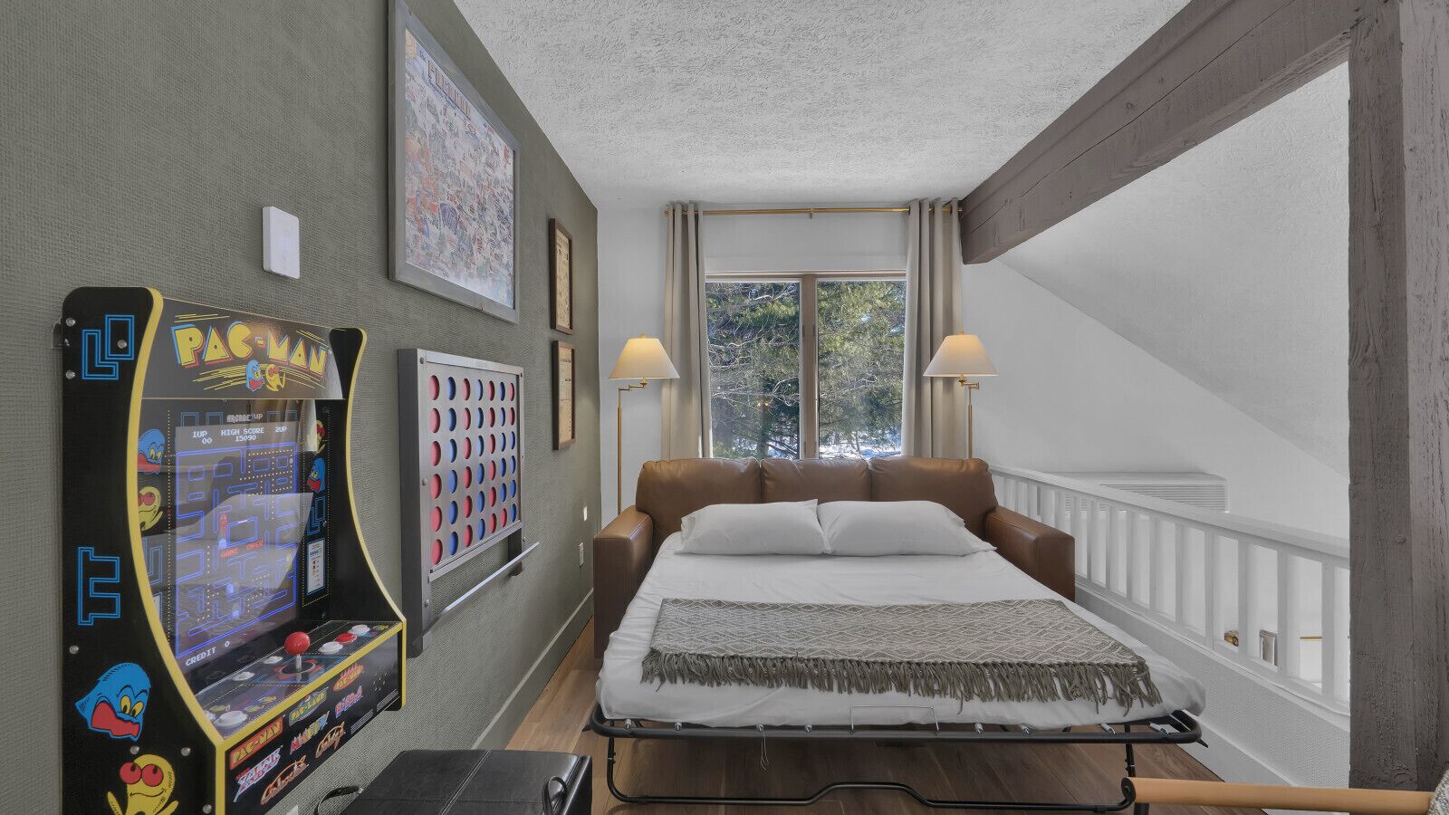 Un dormitorio con cama, máquina arcade Pac-Man y ventana con vistas a árboles.
