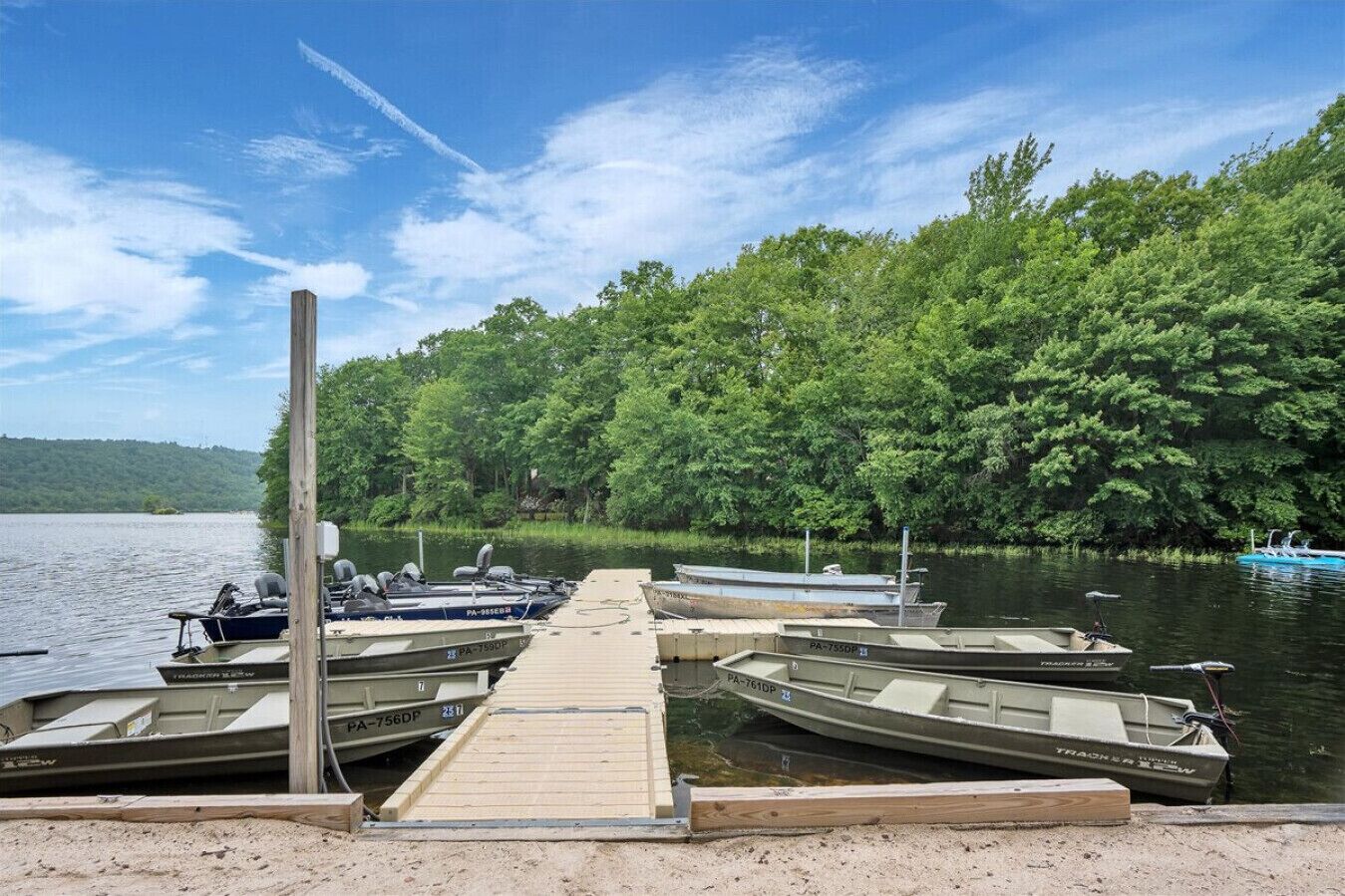 Un muelle con varias embarcaciones amarradas en una orilla arenosa, rodeado de un bosque verde frondoso y un lago tranquilo.