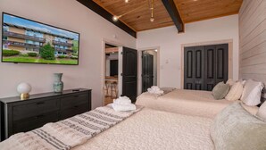 A bedroom with two beds, a TV on the wall, a wooden ceiling, and a door leading to another room. - Sunset Ridge (#338) | Walk to Ski Bike Eat! (Basye)