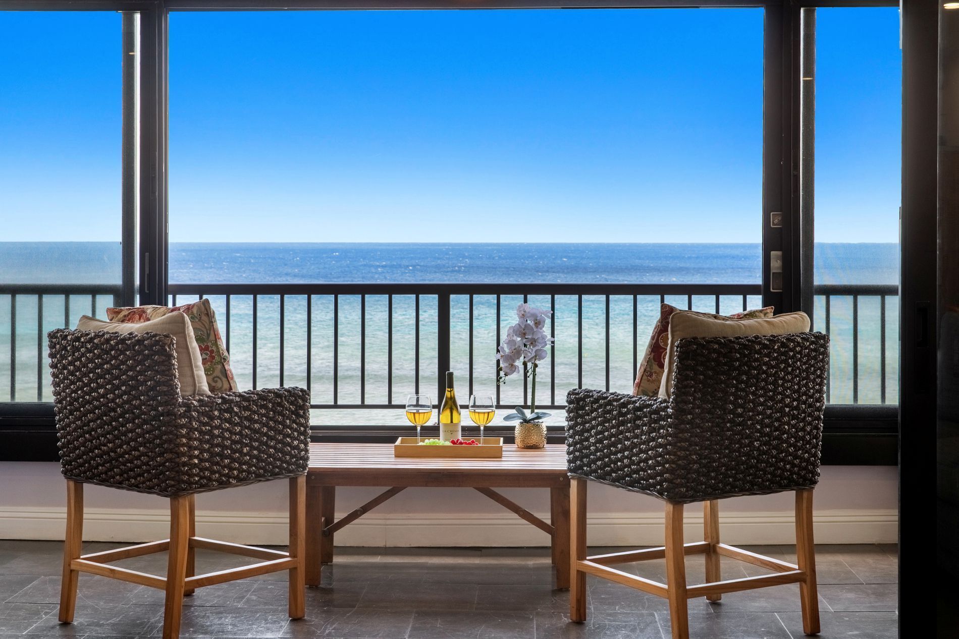 Two wicker chairs with cushions, a wooden table with a bottle and glasses, and a view of the ocean through large windows.
