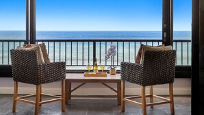 Two wicker chairs with cushions, a wooden table with a bottle and glasses, and a view of the ocean through large windows. - New Listing! Stunning Oceanfront Oasis (Honokowai)