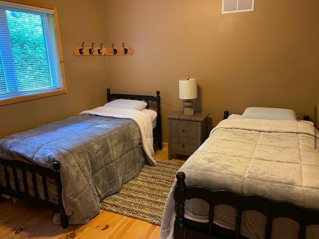 A bedroom with two single beds, a bedside table with a lamp, a window with blinds, and a wall-mounted coat rack.