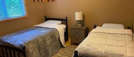 A bedroom with two single beds, a bedside table with a lamp, a window with blinds, and a wall-mounted coat rack.