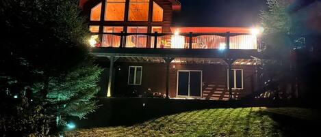 A two-story wooden house with a balcony, illuminated by exterior lights.