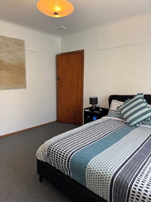 A bedroom with a bed, a nightstand, a lamp, a wooden door, and a wall art piece. - 3BR Airport Stay 10 min to Melbourne Airport (Jacana)