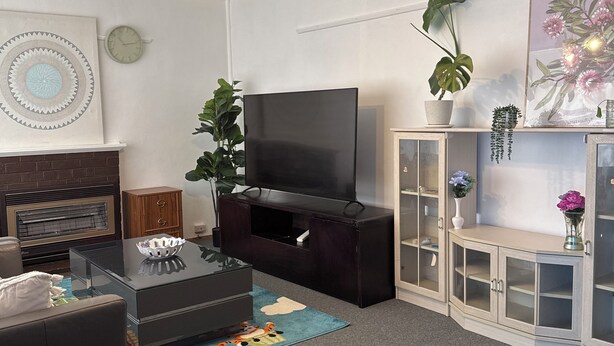A modern living room with a flat-screen TV, a dark entertainment unit, a sofa, a coffee table, and a decorative rug. - 3BR Airport Stay 10 min to Melbourne Airport (Jacana)