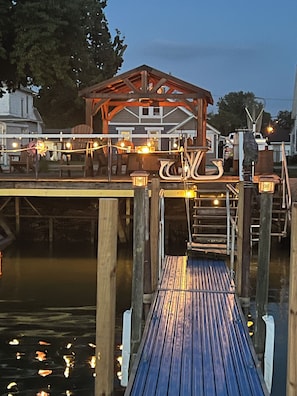 Un muelle de madera con un área de estar cubierta y luces colgantes, que conduce a un edificio con techo inclinado.