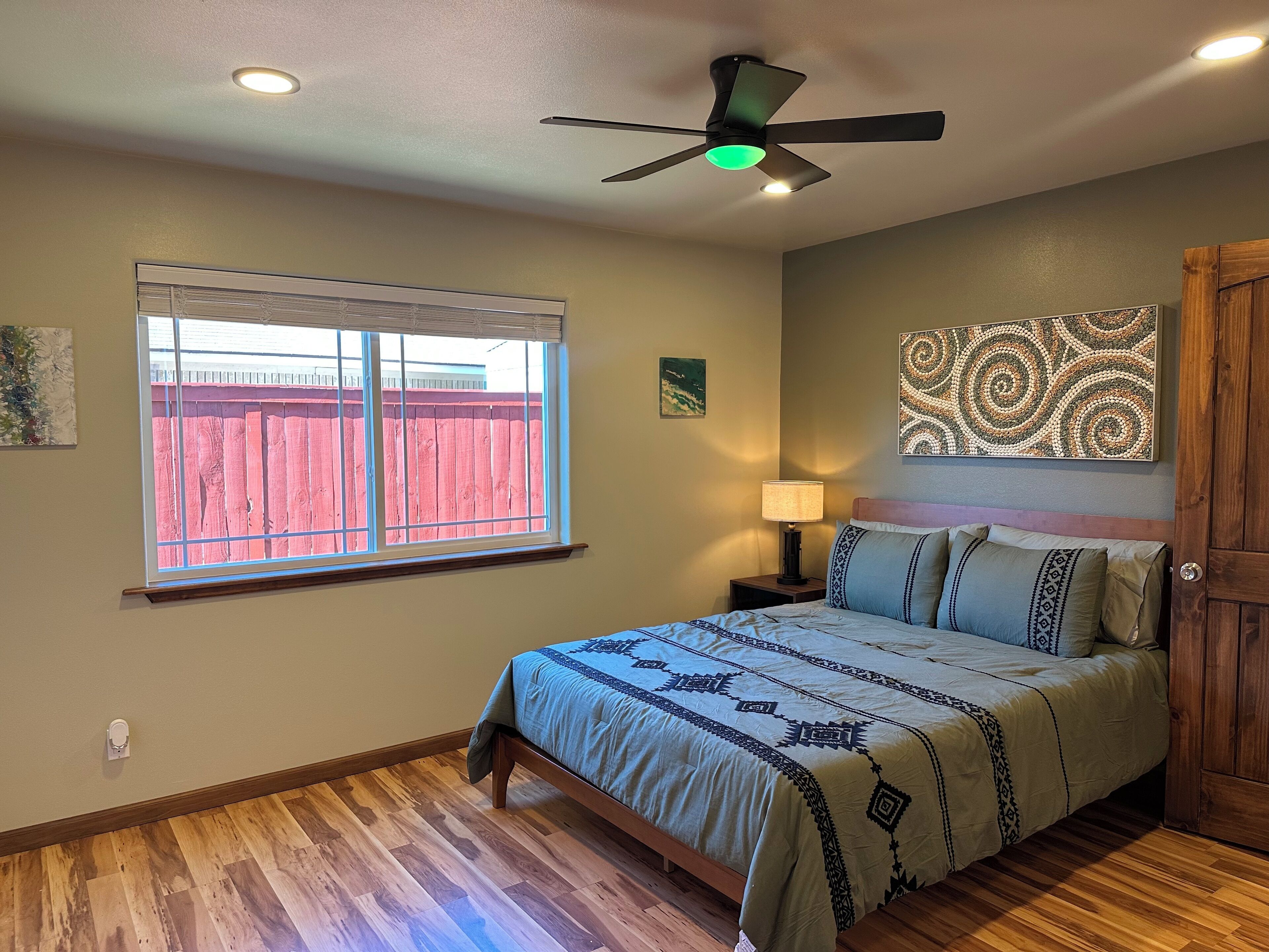 A bedroom with a bed, a nightstand, a ceiling fan, a window with a view of a red fence, and a wooden door.