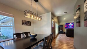 A dining area with a long table, chairs, a bowl of fruit, and a hanging light fixture. - A&C WINDWARD COTTAGE # 2 (Victoria)