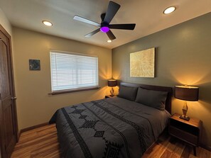 Une chambre à coucher comprenant un lit, un ventilateur de plafond, une fenêtre avec des stores, une table de chevet et une lampe fixée au mur.