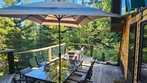 A wooden deck with a glass table and chairs, surrounded by a metal fence and a large umbrella. - Pet friendly off grid chalet tucked away on a mountainside . (Sandpoint)