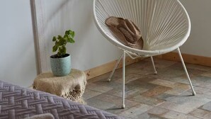 Une pièce comprenant un lit, une chaise en fil de fer blanc, une petite plante en pot et une décoration murale en macramé.