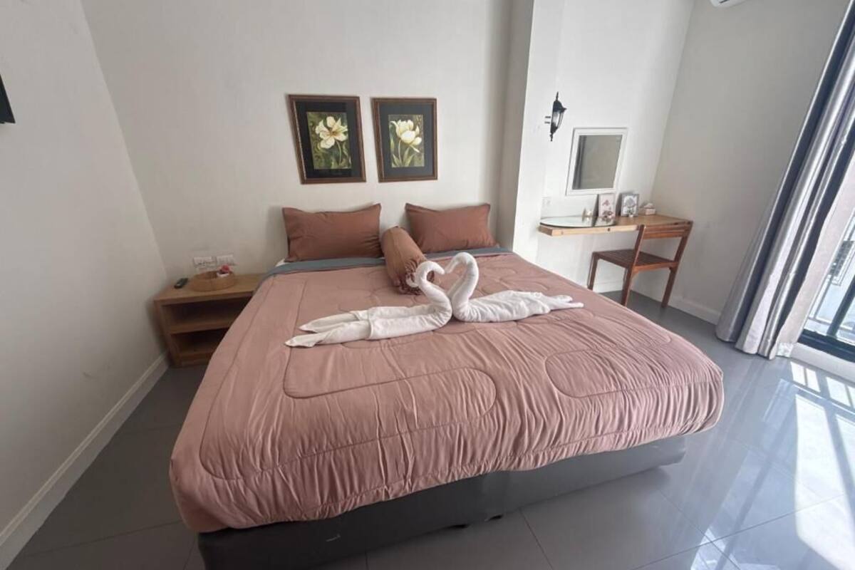 A neatly made bed with a pink bedspread and two pillows, a small wooden nightstand with a lamp, and a desk with a chair.