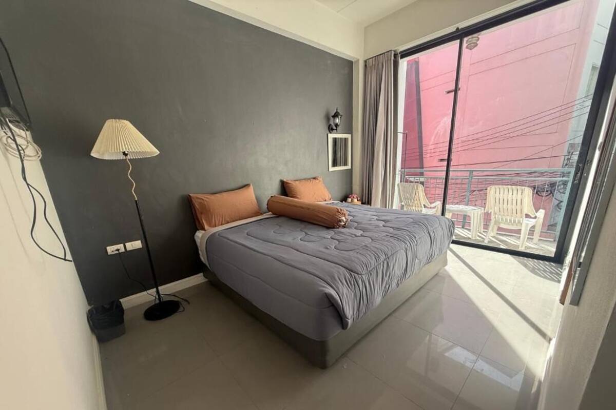 A modern bedroom with a large bed, a standing lamp, and a view of a balcony with chairs.