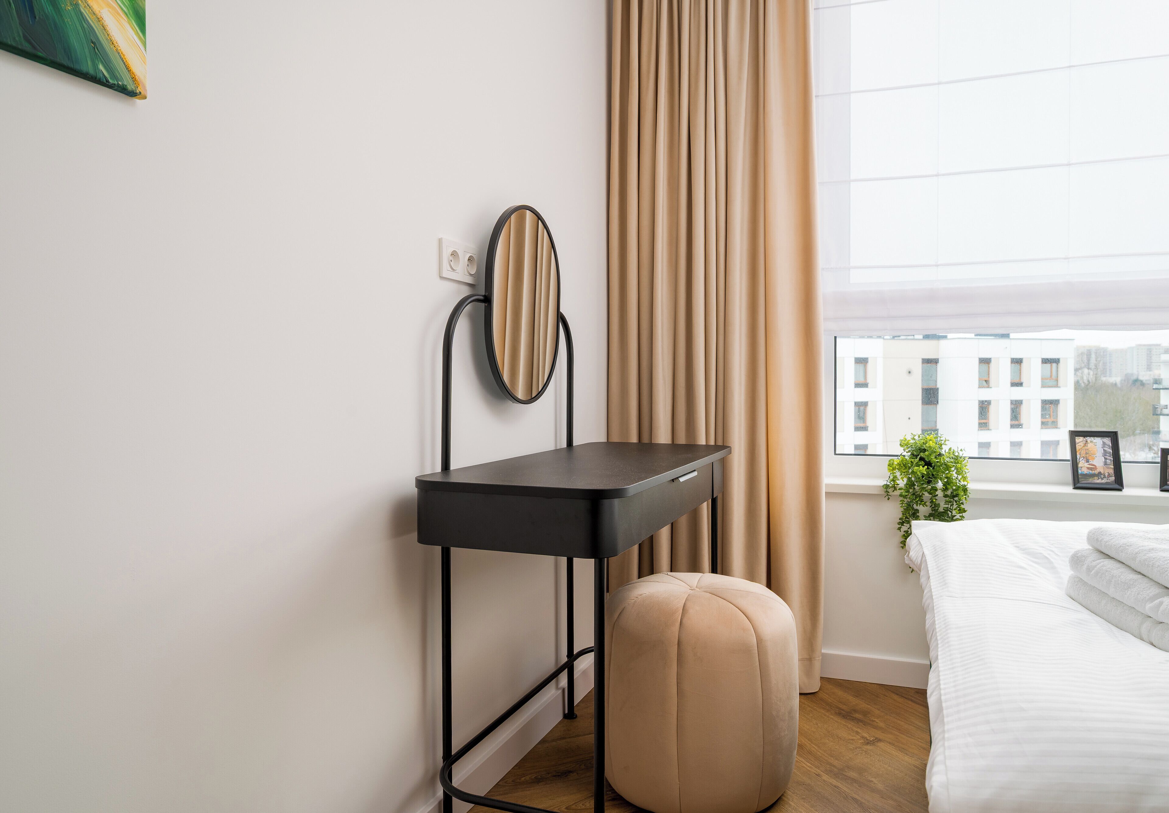 A bedroom with a bed, a bedside table with a mirror, a window with curtains, and a view of buildings outside.