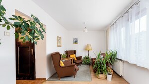 Un salon avec un canapé, un fauteuil et une petite table sur laquelle se trouve une lampe. On y trouve également des plantes en pot et une porte qui donne sur une autre pièce.