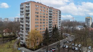 Un immeuble résidentiel de grande hauteur, doté de nombreux balcons, d’un parking avec des voitures et surplombant un ciel dégagé.