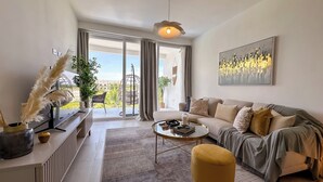 Un salon moderne avec un canapé, un pouf et une table basse. Une grande fenêtre offre une vue sur la verdure environnante.