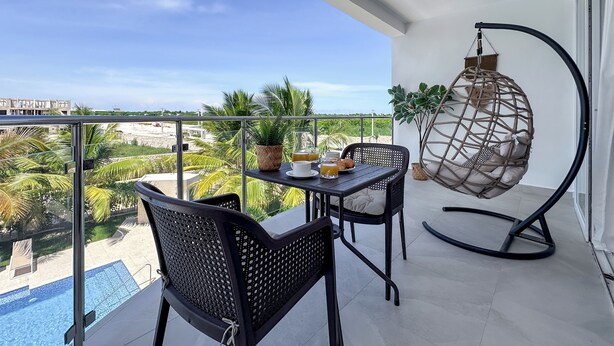 Un balcon avec une table dressée pour deux personnes, un fauteuil suspendu et une vue sur une piscine et des palmiers.