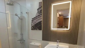 A modern bathroom with a glass-enclosed shower, a toilet, a sink, and a mirror with built-in lighting. - The Gold Leaf Getaway: Stylish 1BR Urban Oasis (Taguig)