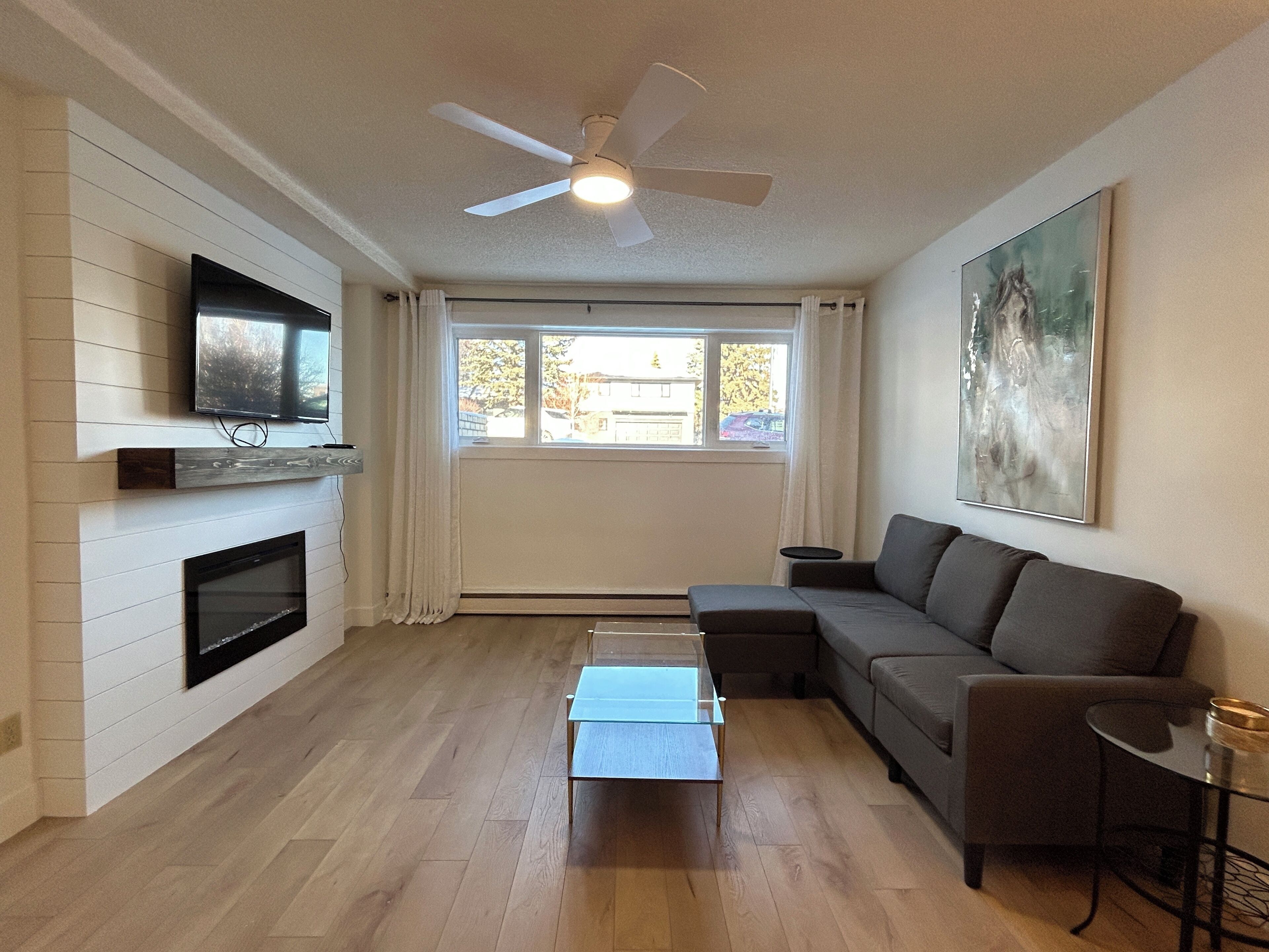 A modern living room with a flat-screen TV mounted on the wall, a fireplace, a ceiling fan, a gray sofa, and a glass coffee table.