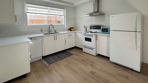 Ett modernt kök med vita skåp, en rostfri köksfläkt, ett vitt kylskåp, en blå mikrovågsugn och ett fönster med persienner.