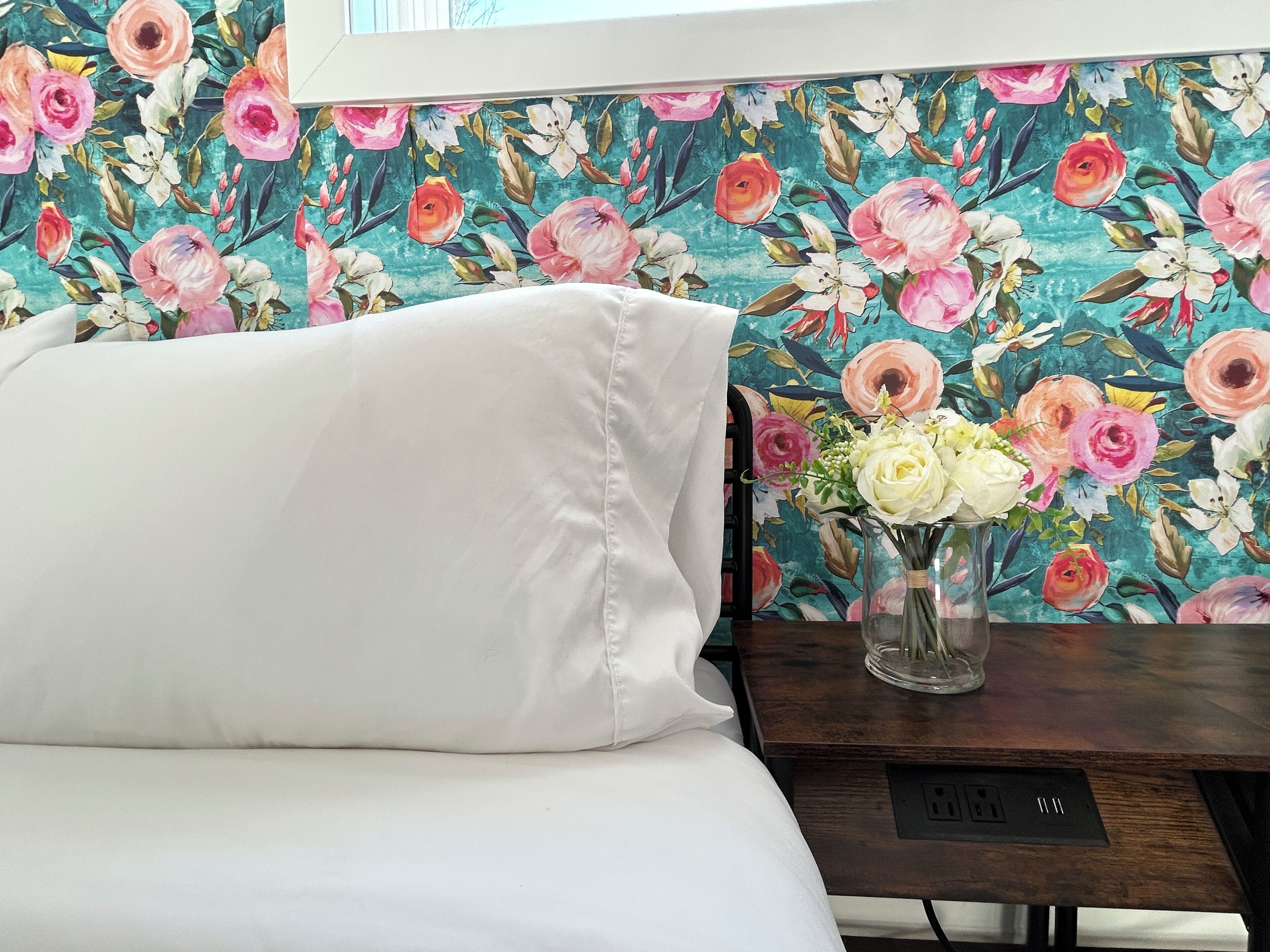Een bed met witte beddengoed, een houten nachtkastje met een bloevas en bloemenbehang.