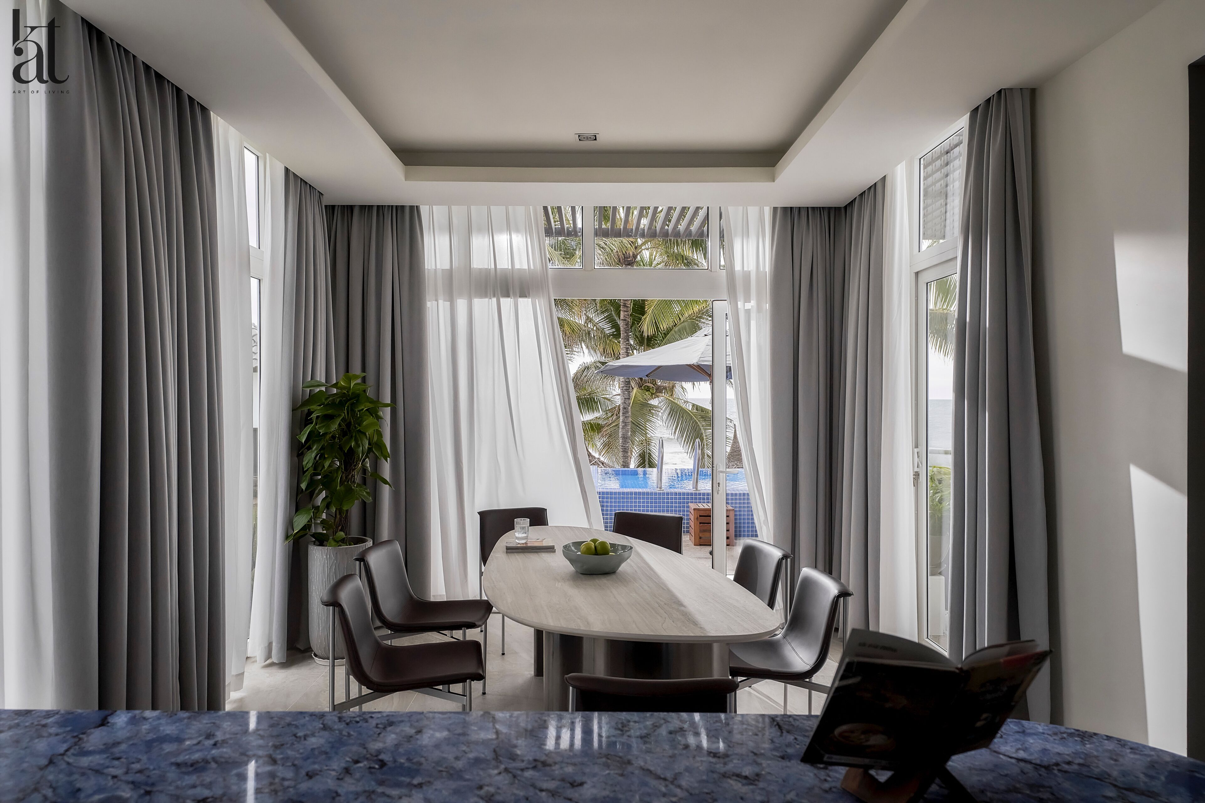 A modern dining area with a marble table, grey curtains, and a view of a pool and palm trees.