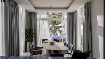 A modern dining area with a marble table, grey curtains, and a view of a pool and palm trees.