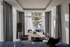 Spiseområde i moderne stil med et marmorbord, grå gardiner og udsigt til et poolområde og palmer.