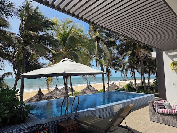An infinity pool with a view of the beach, surrounded by palm trees and a covered seating area.