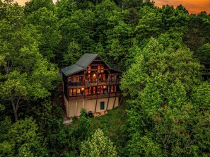A two-story log cabin with a balcony, surrounded by dense forest. - The Overlook - Hot Tub, Game Room, Mountain Views! (Sevierville)