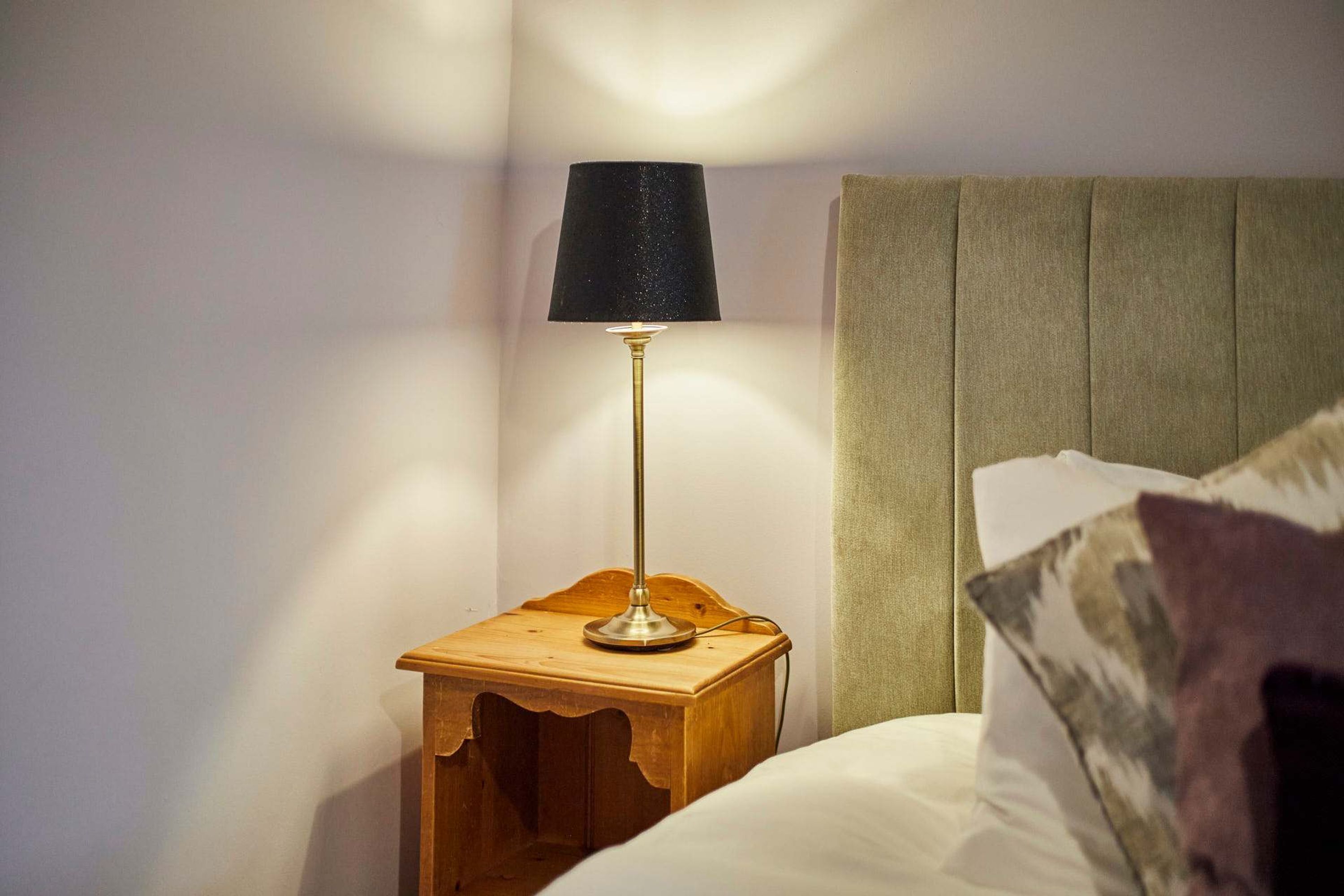 Una mesita de noche de madera con una lámpara, una cama con cabecero y un almohadón.