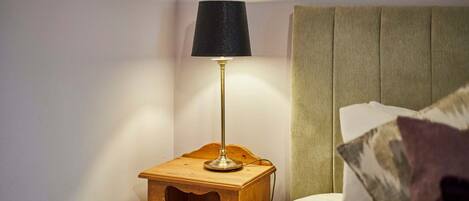 Una mesita de noche de madera con una lámpara, una cama con cabecero y un almohadón.