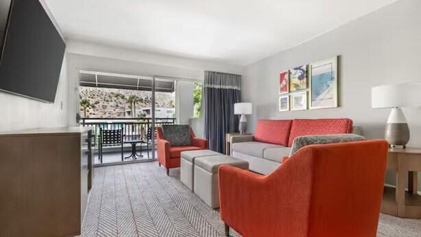 Une chambre d’hôtel avec un canapé, des fauteuils, une télévision, un balcon avec vue et une moquette à motifs.