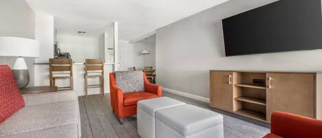 Een moderne woonkamer met een bank, voetenbankje en fauteuil, een flatscreen televisie aan de muur en een houten kast.