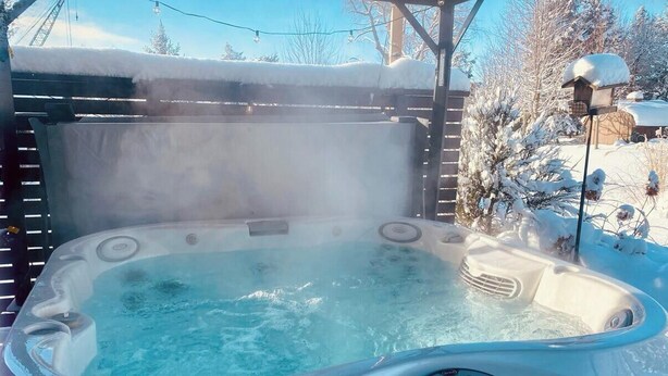 Un jacuzzi al aire libre con vapor ascendiendo, rodeado de árboles cubiertos de nieve y una terraza de madera.