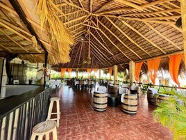 Un espacio al aire libre techado con vigas de madera, mesas, sillas y una zona de bar.