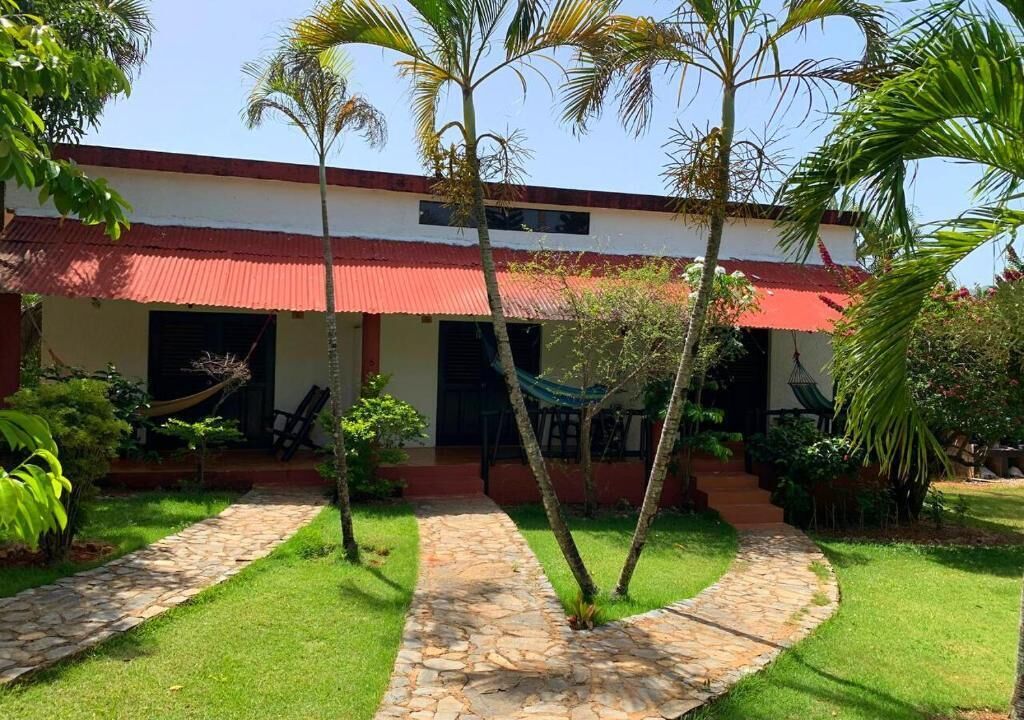 Un edificio con techo rojo y paredes blancas, rodeado de un jardín bien cuidado con senderos de piedra y palmeras.
