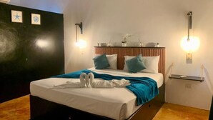 Een net opgemaakt bed met een blauwe deken en witte kussens, een nachtkastje met een lamp, en wandlampen.