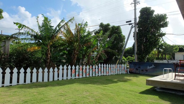 A grassy yard with a white picket fence, palm trees, and a mural on a wall. - Heath Miyakojima (Miyakojima)
