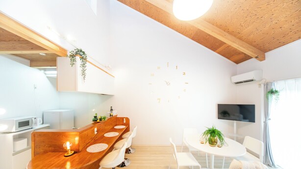 A modern kitchen with wooden accents, a central island with stools, a dining area with a round table and chairs, and a wall-mounted TV. - Miraie Yokkaichi Hinaganishi (Yokkaichi)