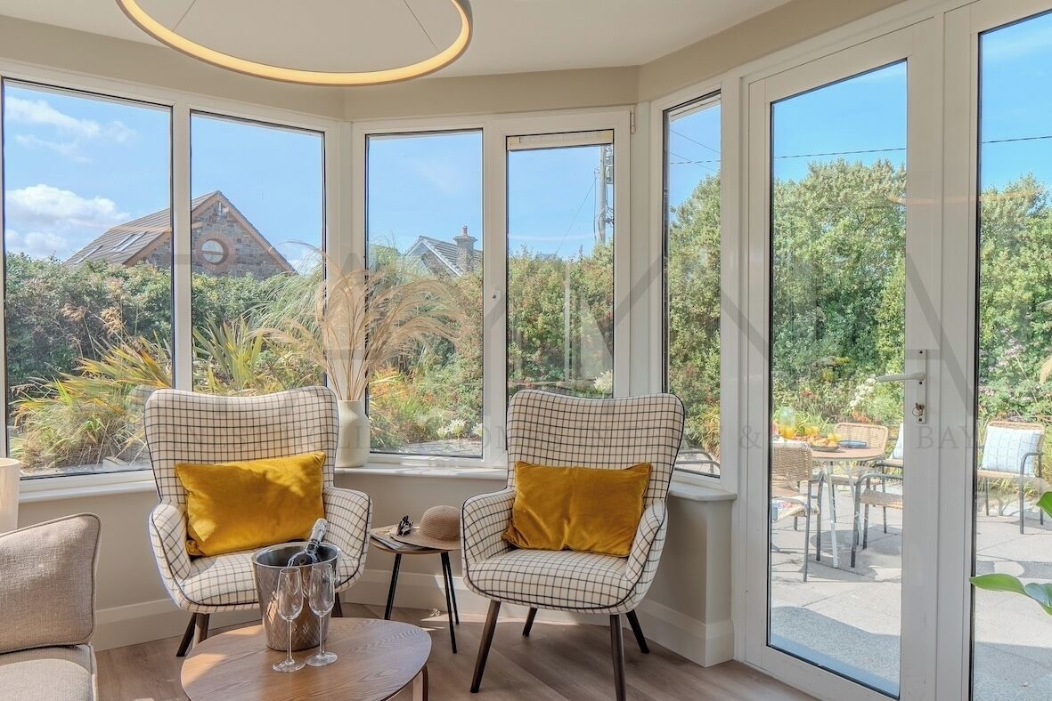 A room with large windows, two checkered chairs with yellow cushions, a small table with a glass of wine, and a view of a house and greenery outside.