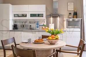 A modern kitchen with white cabinets, a stainless steel range hood, and a built-in oven. There is a dining table with chairs, a bowl of fruit, and a croissant on the table. - Margot - 1 bedroom apartment (Nice)